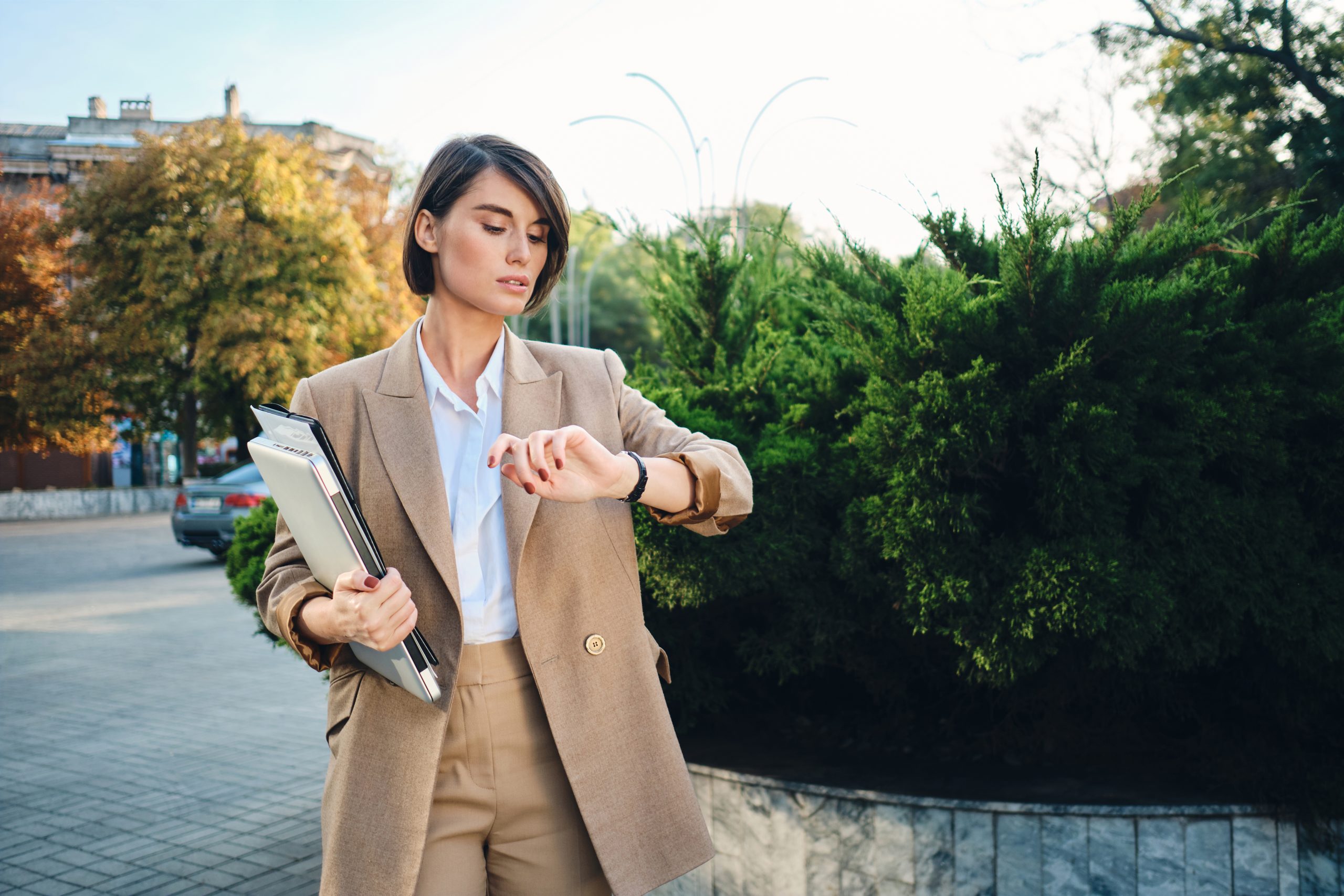 Young confident businesswoman in beige suit standing with laptop watching time on watch outdoor